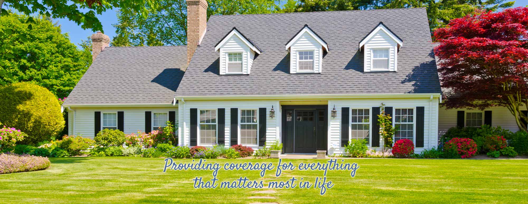 Exterior of a large family home with white siding, black shutters, flowering shrubs, and a well manicured lawn.